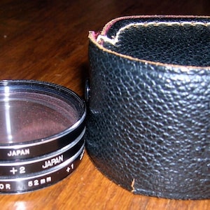 May include: A set of three black camera lens filters with the text "Soligor 52mm +2 Japan" and "52mm +5 Japan" printed on them. The filters are in a black leather case.