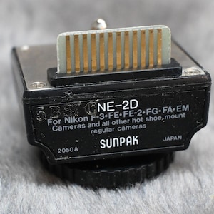 May include: A black Sunpak NE-2D hot shoe adapter for Nikon cameras. The adapter has a silver and gold connector and the text "SUNPAK" and "JAPAN" are visible. The adapter is designed for Nikon F-3, FE, FE-2, FG, FA-EM cameras.