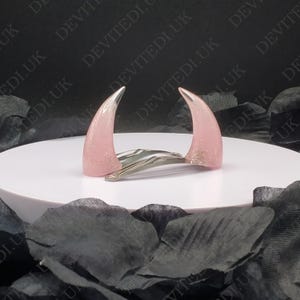 May include: Pink devil horn hair clips with silver tips, set on a silver hair clip. The horns have glitter and are set against a black backdrop with the text "DEVITEDI.UK" repeated.