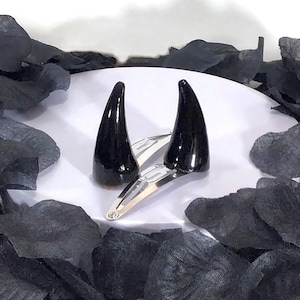 May include: A pair of black, glossy devil horn hair clips. The horns are mounted on silver-colored metal clips. The background features black, petal-shaped decorations on a white surface, creating a contrasting aesthetic.
