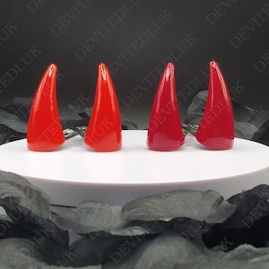 May include: Four glossy red devil horn-shaped nail art accessories. Each horn is a different shade of red, from bright to deep. They are displayed on a white surface against a dark background with the text "DEVITEDI.UK".