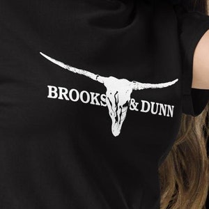 May include: Black t-shirt with a white bull skull graphic and the text "BROOKS & DUNN". The shirt is made of a soft material and is paired with blue jeans. The design is centered on the chest.