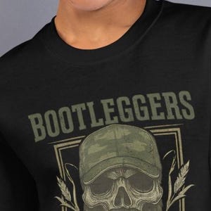 May include: Black sweatshirt featuring a skull wearing a camouflage baseball cap and a long beard. The word "BOOTLEGGERS" is printed above the skull in a rectangular frame. The design includes wheat stalks on either side of the skull.