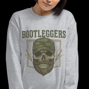 May include: A light gray sweatshirt with the word "BOOTLEGGERS" above a skull wearing a camouflage hat and a beard. The skull graphic is framed with a rectangle and wheat stalks.