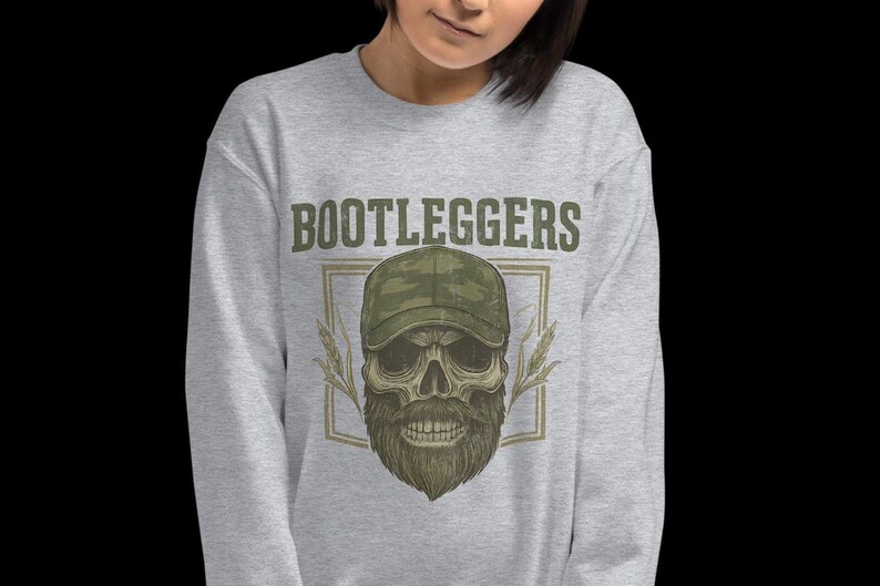 May include: A light gray sweatshirt with the word "BOOTLEGGERS" above a skull wearing a camouflage hat and a beard. The skull graphic is framed with a rectangle and wheat stalks.