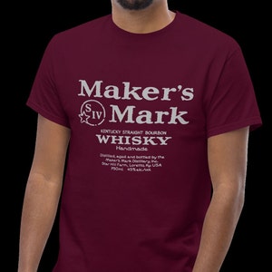 May include: A maroon t-shirt with a white graphic of the Maker's Mark whiskey label. The label includes the text "Maker's Mark", "Kentucky Straight Bourbon", "Whisky", "Handmade", and details about the distillery and alcohol content.
