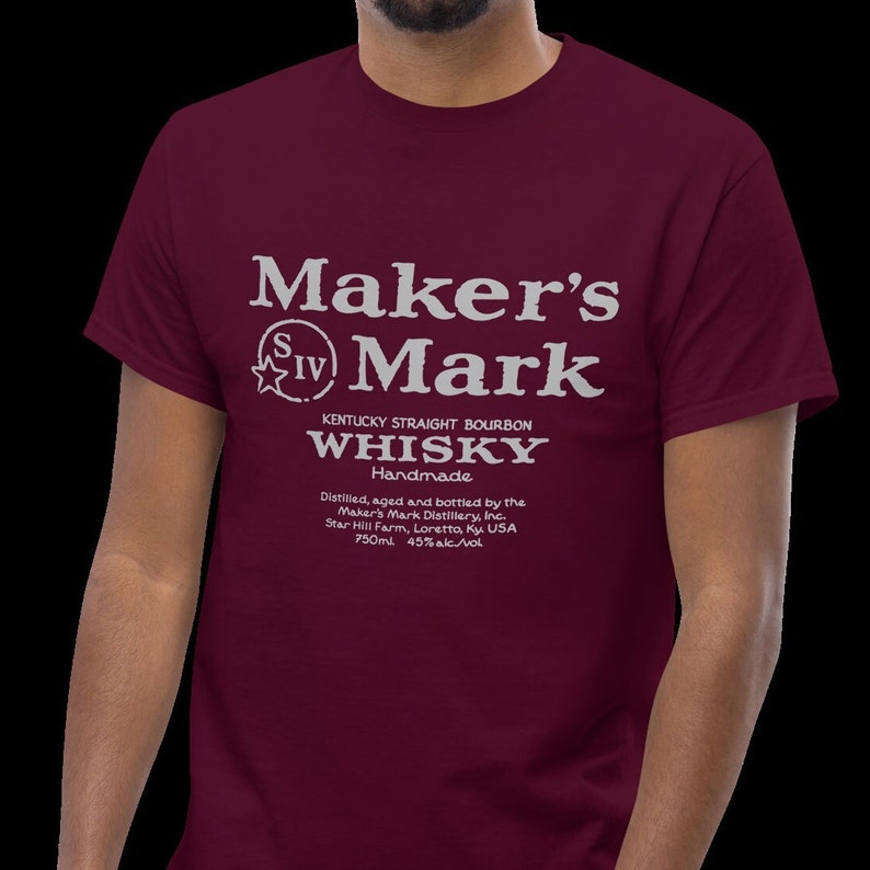 May include: A maroon t-shirt with a white graphic of the Maker's Mark whiskey label. The label includes the text "Maker's Mark", "Kentucky Straight Bourbon", "Whisky", "Handmade", and details about the distillery and alcohol content.