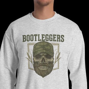 May include: Light gray sweatshirt with the word "BOOTLEGGERS" above a skull wearing a camouflage cap and a full beard. The skull is framed by a rectangular border with wheat stalks on either side. The design is in shades of green, brown, and tan.