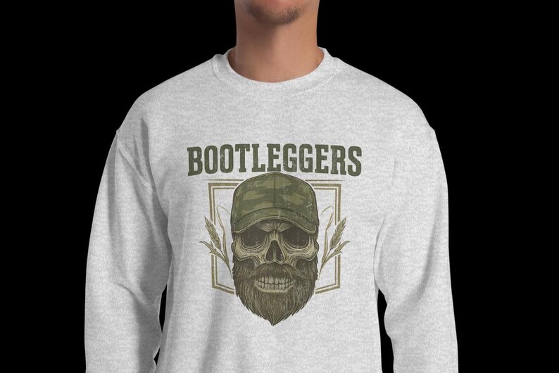 May include: Light gray sweatshirt with the word "BOOTLEGGERS" above a skull wearing a camouflage cap and a full beard. The skull is framed by a rectangular border with wheat stalks on either side. The design is in shades of green, brown, and tan.