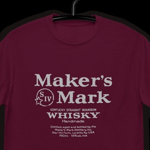 May include: A maroon t-shirt with a white graphic of Maker's Mark Kentucky Straight Bourbon Whisky. The graphic includes the text "Maker's Mark", "SIV", "Handmade", "Distilled, aged and bottled by the Maker's Mark Distillery, Inc.", "Star Hill Farm, Loretto, Ky. USA", "750ml", and "45% alc./vol".