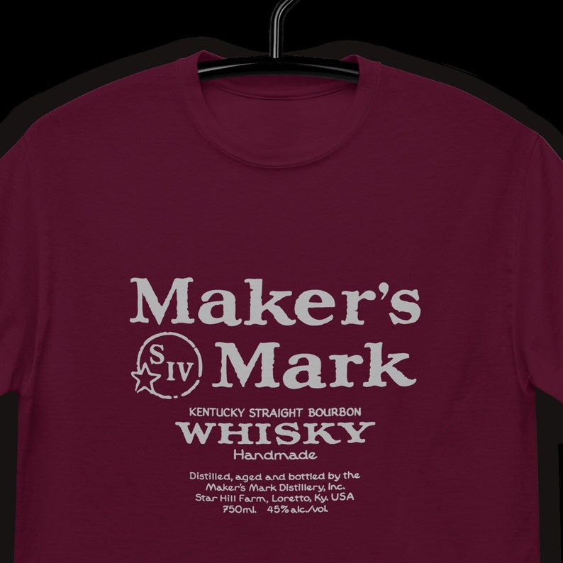 May include: A maroon t-shirt with a white graphic of Maker's Mark Kentucky Straight Bourbon Whisky. The graphic includes the text "Maker's Mark", "SIV", "Handmade", "Distilled, aged and bottled by the Maker's Mark Distillery, Inc.", "Star Hill Farm, Loretto, Ky. USA", "750ml", and "45% alc./vol".