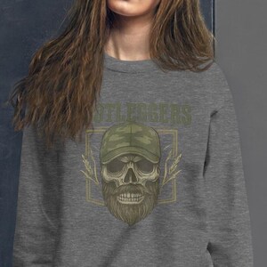 May include: A gray sweatshirt featuring a skull design with a beard and a camouflage baseball cap. The word "STILLEGERS" is printed above the skull in a green, distressed font. The design is framed by a gold-colored rectangle.
