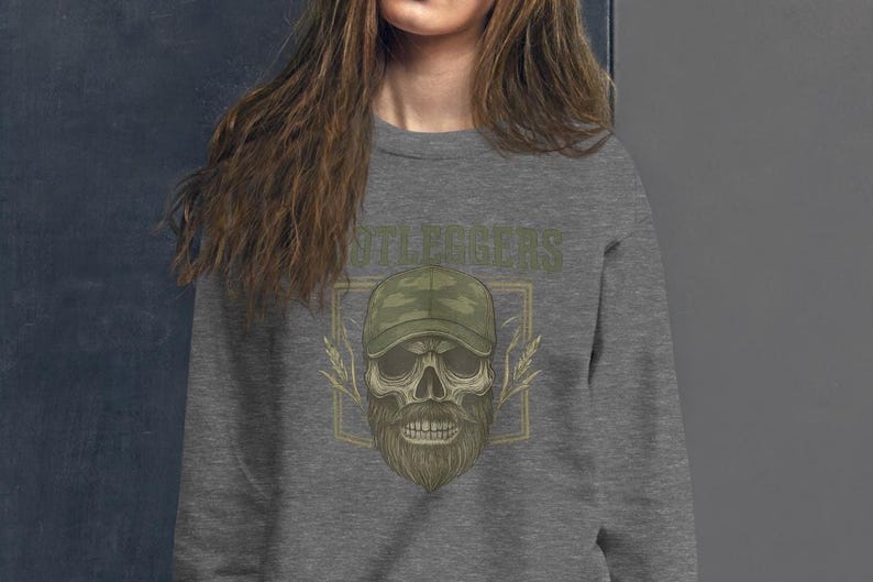 May include: A gray sweatshirt featuring a skull design with a beard and a camouflage baseball cap. The word "STILLEGERS" is printed above the skull in a green, distressed font. The design is framed by a gold-colored rectangle.