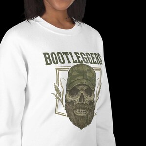May include: White sweatshirt featuring the word "BOOTLEGGERS" above a skull wearing a camouflage baseball cap and a long beard. The design includes wheat stalks and a rectangular border.