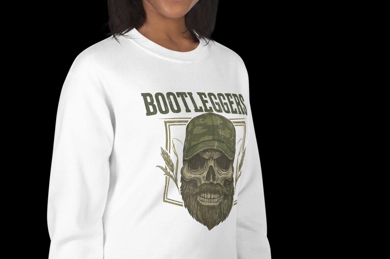 May include: White sweatshirt featuring the word "BOOTLEGGERS" above a skull wearing a camouflage baseball cap and a long beard. The design includes wheat stalks and a rectangular border.