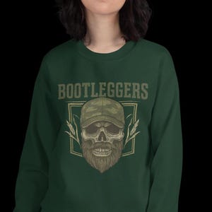 May include: Dark green sweatshirt with the word "BOOTLEGGERS" above a skull wearing a camouflage baseball cap and a long beard. The skull is framed by a square with wheat stalks on either side.