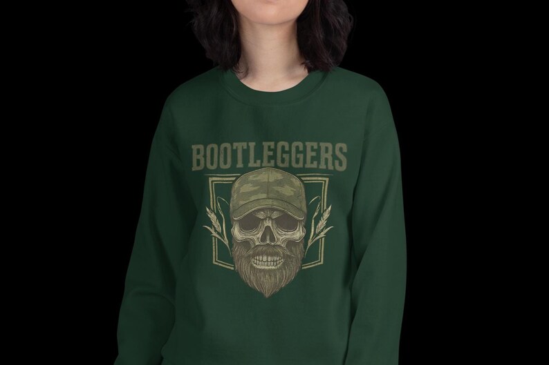May include: Dark green sweatshirt with the word "BOOTLEGGERS" above a skull wearing a camouflage baseball cap and a long beard. The skull is framed by a square with wheat stalks on either side.