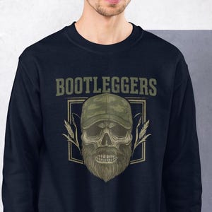 May include: Navy blue sweatshirt featuring a skull wearing a camouflage baseball cap and a beard. The word "BOOTLEGGERS" is printed above the skull in a green, vintage font. The design includes wheat stalks on either side of the skull.