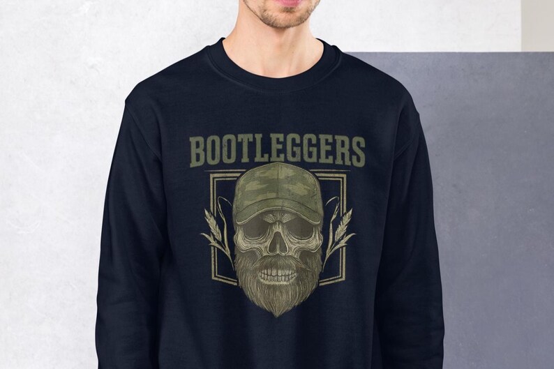 May include: Navy blue sweatshirt featuring a skull wearing a camouflage baseball cap and a beard. The word "BOOTLEGGERS" is printed above the skull in a green, vintage font. The design includes wheat stalks on either side of the skull.