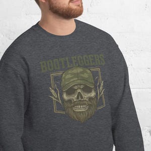 May include: Dark gray sweatshirt featuring a skull wearing a camouflage baseball cap and a long beard. The word "BOOTLEGGERS" is printed above the skull in a green, vintage-style font. The design is enclosed in a rectangular frame.