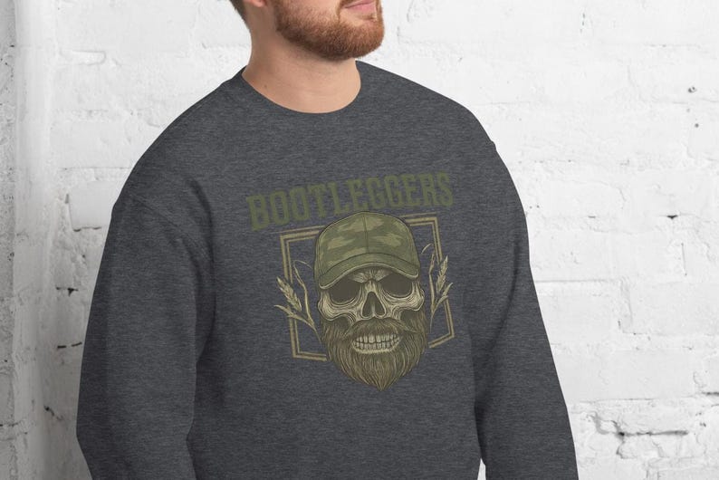 May include: Dark gray sweatshirt featuring a skull wearing a camouflage baseball cap and a long beard. The word "BOOTLEGGERS" is printed above the skull in a green, vintage-style font. The design is enclosed in a rectangular frame.