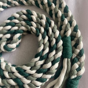 May include: A thick, braided rope made of intertwined strands in shades of forest green, sage green, and off-white. The rope is coiled in a circular pattern, featuring a tightly wrapped green cord section that secures the ends into a decorative tassel.