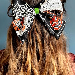 May include: A large fabric hair bow featuring a black background with white spiderwebs, orange and black buffalo plaid pumpkins, and text that reads "WITCH way to the house". The centre of the bow is cinched with a bright green fabric knot.