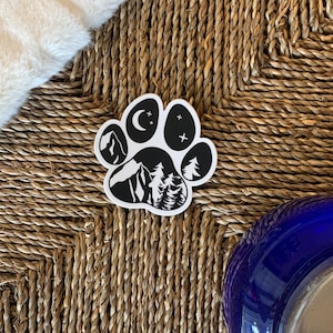May include: Black and white paw print sticker with a mountain, trees, moon, and stars inside. The sticker is on a woven surface.