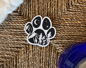 May include: Black and white paw print sticker with a mountain, trees, moon, and stars inside. The sticker is on a woven surface.