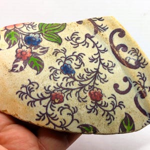 Puede incluir: Fragmento de cerámica antigua con un diseño floral. El diseño presenta flores rojas, azules y rosas con hojas verdes y enredaderas marrón oscuro sobre un fondo crema. La pieza tiene una apariencia desgastada y envejecida.