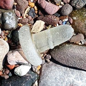 May include: A translucent, frosted glass bottle stopper with a wide, rounded top and a tapered, cylindrical base featuring a raised horizontal band. The object has a smooth, weathered texture and is positioned among various smooth, multi-colored stones.