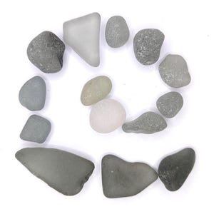 May include: A collection of sea glass pieces in various shapes and shades of gray, arranged in a circular pattern against a white background. The pieces vary in size and texture, with some appearing smooth and others more rough.