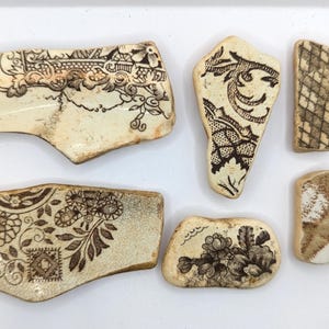 May include: A selection of antique ceramic fragments, featuring brown floral and geometric patterns on a cream base. The pieces, of varying sizes and shapes, display intricate designs from vintage tableware.