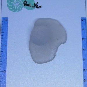 Puede incluir: Una pieza de vidrio de mar lisa y blanca, con una forma ligeramente irregular. Mide aproximadamente 5 cm de largo.