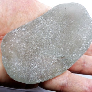 May include: A smooth, grey sea glass stone with a slightly green tint. The stone has a rough, textured surface and is slightly translucent.