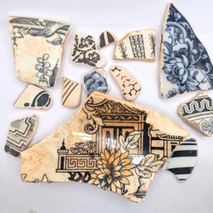May include: A collection of antique ceramic shards with various black and white patterns. Designs include floral motifs, geometric shapes, and architectural elements. The fragments have a weathered, aged appearance, suggesting they are vintage pieces.