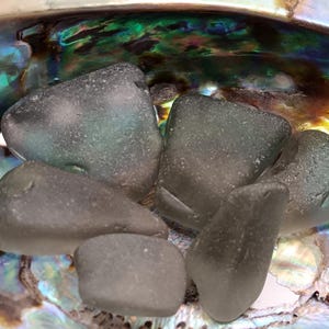 May include: Five pieces of sea glass in various shades of green and gray, resting in a mother-of-pearl shell.