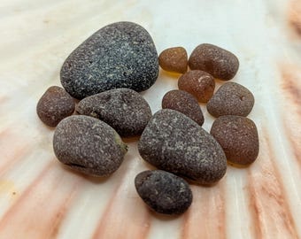 Könnte beinhalten: Eine Ansammlung glatter, abgerundeter Steine in Grau- und Brauntönen, die auf einer hellen, strukturierten Oberfläche liegen. Die Steine variieren in Größe und Form, mit einigen kleineren, bernsteinfarbenen Stücken dazwischen. Das Arrangement deutet auf ein natürliches, organisches Thema hin.
