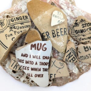 Puede incluir: Fragmentos de cerámica antiguos con texto negro descolorido sobre fondo beige. Los fragmentos muestran palabras como "Ginger", "Beer" y "Mug". Las piezas están dispuestas sobre una superficie texturizada de color blanquecino.