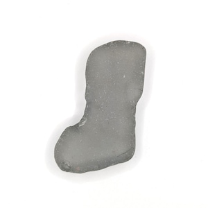 May include: A gray, irregularly shaped piece of sea glass, resembling a stocking, against a white background. The glass has a smooth, frosted appearance and a slightly translucent quality. The object is likely a decorative item or a component of jewelry.