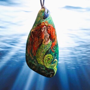 May include: A green stone pendant with a painted mermaid design. The mermaid has red hair, a crown, and a green tail. The pendant is hanging from a blue cord.