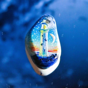 May include: A white stone painted with a lighthouse scene. The lighthouse is yellow and white with a blue roof. The sky is blue with white clouds and a crescent moon. The water is blue and there is a sunset in the background.