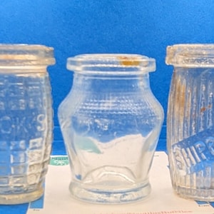 Puede incluir: Tres tarros de vidrio antiguos de diferentes formas y tamaños. El tarro de la izquierda tiene la palabra "PECK'S" en relieve. El tarro del medio es pequeño y tiene forma de reloj de arena. El tarro de la derecha tiene la palabra "SHIPPAM" en una etiqueta azul.