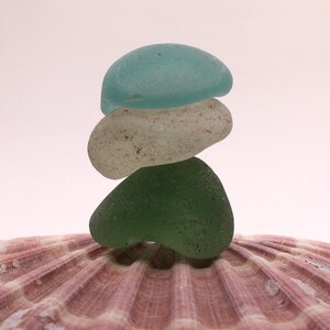 May include: A stack of three sea glass stones, one light blue, one white, and one green, balanced on a pink seashell.
