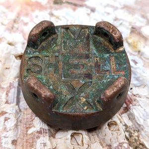 Puede incluir: Objeto metálico de color bronce desgastado con la palabra "SHELL" grabada. Tiene forma circular con cuatro protuberancias redondeadas y un diseño en forma de cruz. Muestra signos de antigüedad.