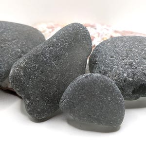 May include: A collection of smooth, gray beach stones of varying sizes. The stones have a textured surface and are arranged on a white background. The stones are a natural, neutral color.