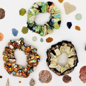 Puede incluir: Tres scrunchies de pelo coloridos con diferentes patrones. El primer scrunchie es un patrón floral verde, azul y blanco. El segundo scrunchie es un patrón marrón, blanco y dorado con un diseño de concha. El tercer scrunchie es un patrón blanco y negro con un diseño floral.