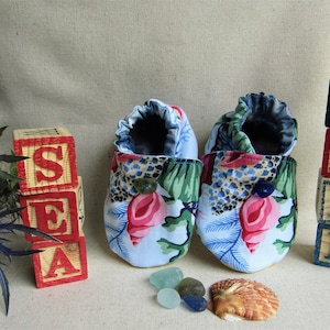 May include: A pair of baby shoes with a floral print featuring pink, blue, and green colors. The shoes are made of fabric and have a button on the top. The shoes are next to a stack of wooden blocks that spell out "SEA" and another stack that spells out "SHELL".