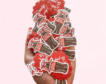 May include: A collection of die-cut stickers featuring brown glass soda bottles with red ribbons and the text "Cola" in white script. Some stickers are circular with scalloped edges and the text "Ready to pop!" in white script on a red background.