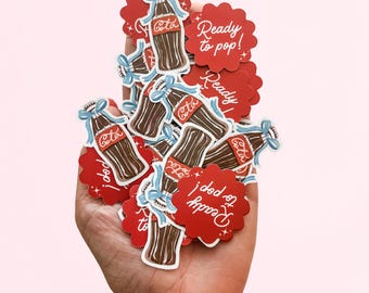 Puede incluir: Una colección de pegatinas troqueladas con botellas de refresco de cristal marrón con lazos azul claro y pegatinas circulares festoneadas rojas con el texto blanco Ready to pop! La palabra Cola está escrita en blanco sobre el cristal.